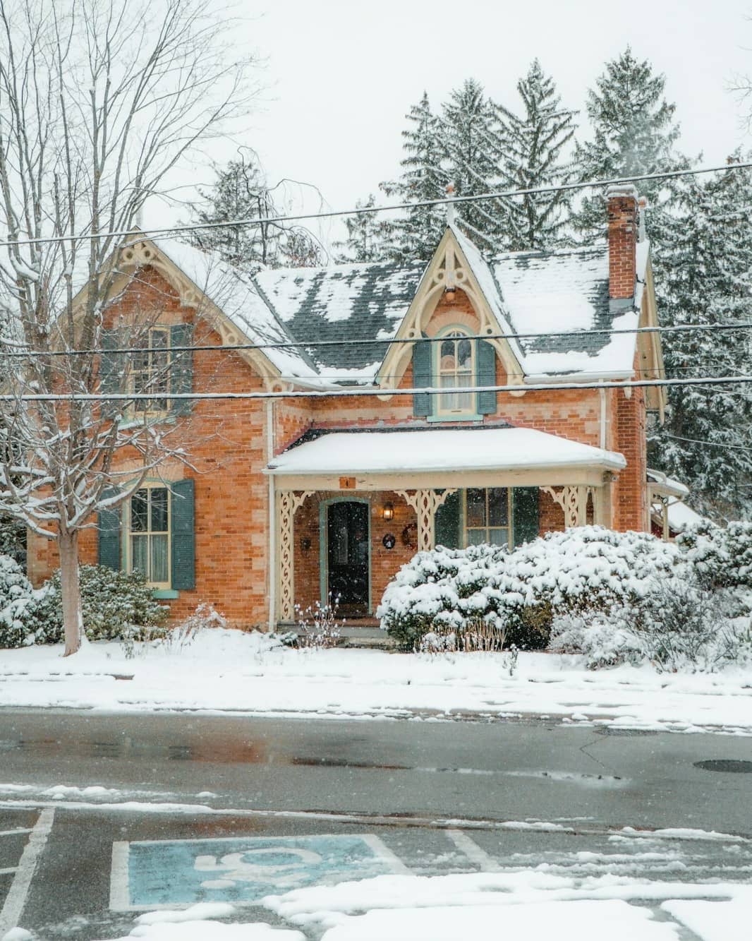 A red-brick Ontario Gothic house with gingerbread trim under fresh snow, its porch and evergreens dusted white.