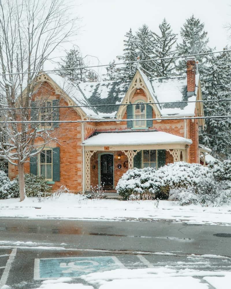 A red-brick Ontario Gothic house with gingerbread trim under fresh snow, its porch and evergreens dusted white.