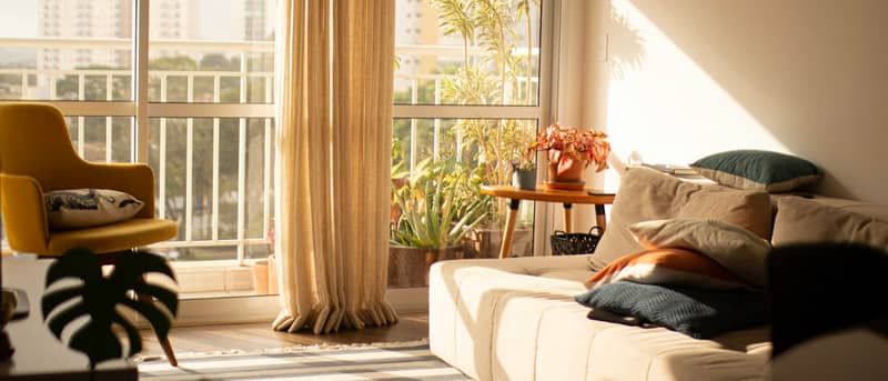A sun-filled apartment living room with a linen sofa, plants, a mustard chair and long window shadows.